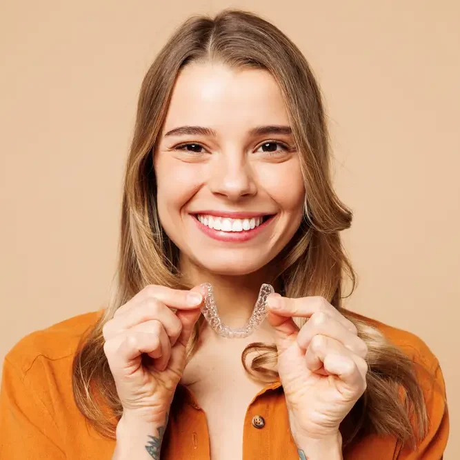 young lady wearing orange top smiling with straight white teeth while holding Invisalign aligners